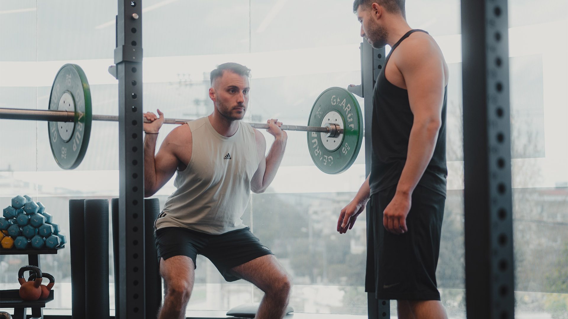 Icon Fitness | άντρας που κάνει κάθισμα με μπάρα και γυμναστής δίπλα του