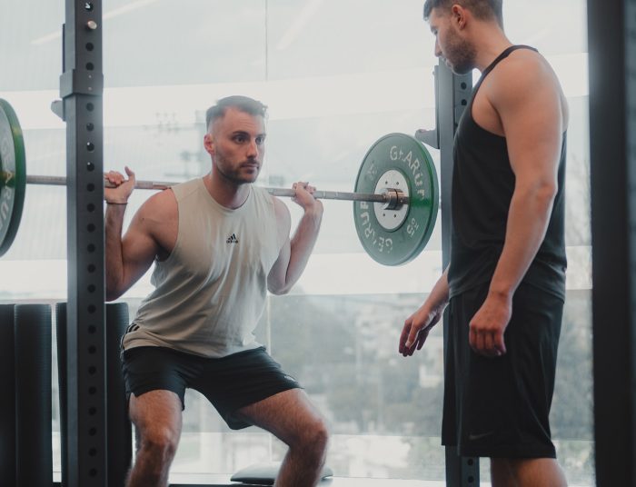Icon Fitness | άντρας που κάνει κάθισμα με μπάρα και γυμναστής δίπλα του